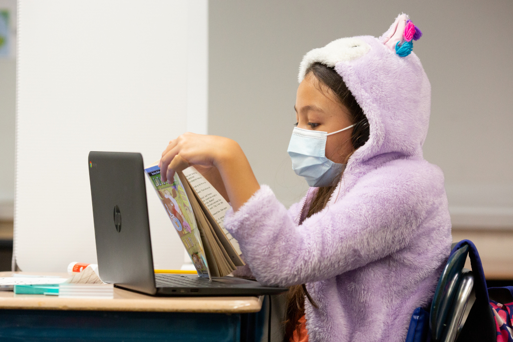 En la imagen puedes ver una niña sentada en una silla, leyendo un libro de papel, que está delante de un portátil abierto