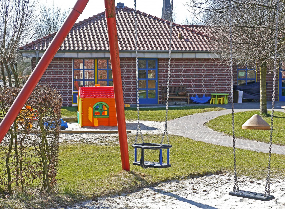 En la imagen puedes ver una zona de juegos al aire libre, con árboles alrededor, donde vemos columpios y una casita de juguete. Al fondo se ve un edificio que podría ser un colegio