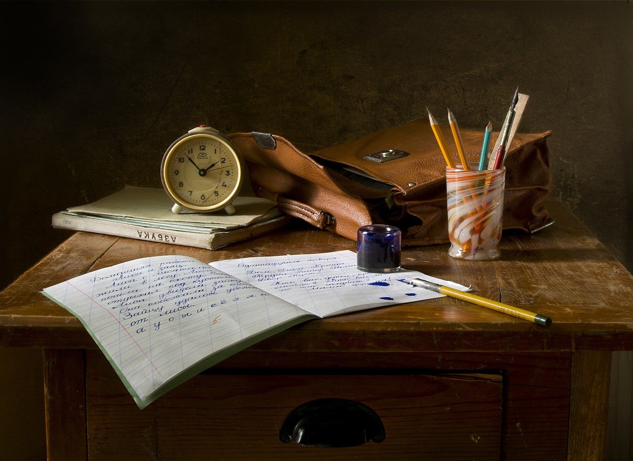 Bodegón de escritura en el que hay sobre una mesa una cartera, un lapicero con tres lápices y una pluma, una libreta abierta, una pluma, un tintero, un libro y un reloj de mesita